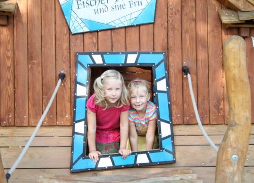 Zwei Mädchen gucken aus einem Holzhaus des Niedrigseilgarten heraus. Der Rahmen ist blau und weiß.