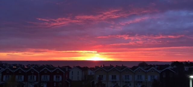 Über den kleinen Häuschen des Resorts geht die Sonne rosafarben unter. Darunter das Meer.