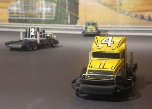 Drei nummerierte Super Trucks stehen und warten auf ihre nächste Fahrt. An der Wand ist ein Truck.