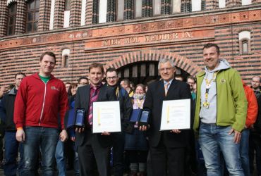 Zwei Männer mit einer Urkunde und einem Präsent stehen vor einer Menge von Menschen.