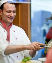 Ein Mann in HANSA-PARK Arbeitskleidung bereitet Pasta zu. Er lächelt.