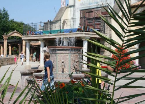 Ein Junge läuft um den sprechenden Brunnen. Oben aus dem Brunnen sprudelt aus den Fontänen Wasser.