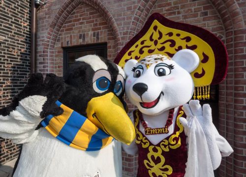 Die Maskottchen Emmi und Pingi stehen vor einem Gebäude. Sie winken beide.