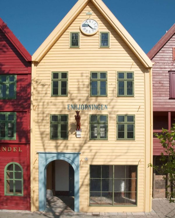 Das Gebäude Enhjorningen aus Norwegen. Es ist gelb mit grünen Fenstern. Oben ist eine Uhr befestigt.