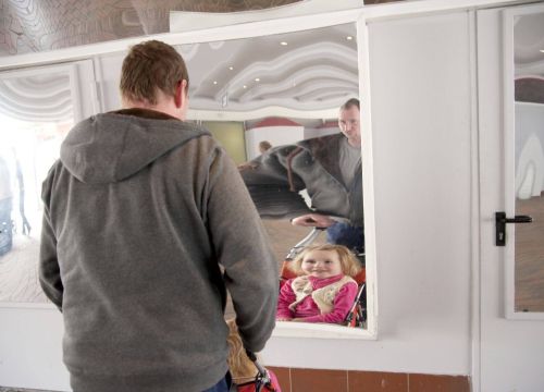 Ein Mann steht mit einem Mädchen im Kinderbuggy in der Spiegelgalerie. Sie stehen vor einem Spiegel.