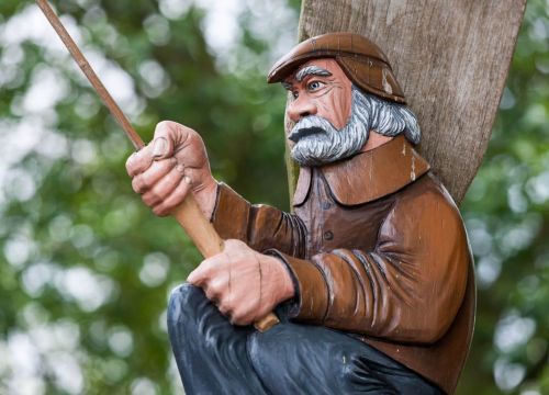 Die Holzfigur des Niedrigseilgarten ist ein alter Mann. Dieser hat eine Mütze auf und eine Angel.