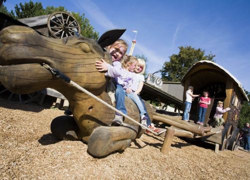 Drei Mädchen sitzen auf einem liegenden Pferd. Auf der Postkutsche dahinter stehen drei Mädchen.
