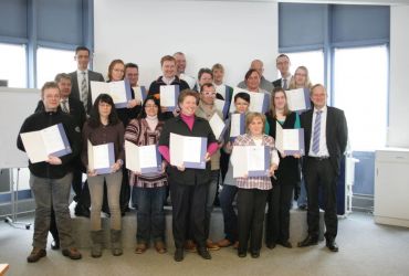 Menschen in drei Reihen. Sie halten alle eine Urkunde in den Händen. Sie schauen in die Kamera.