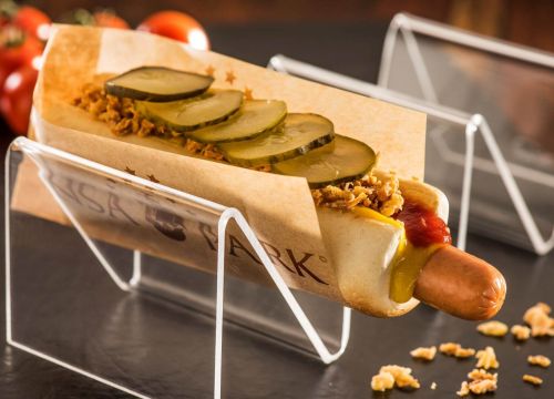 Hotdog des Kiosk Filling Station. Röstzwiebeln im Vorder- und Tomaten im Hintergrund zu sehen.