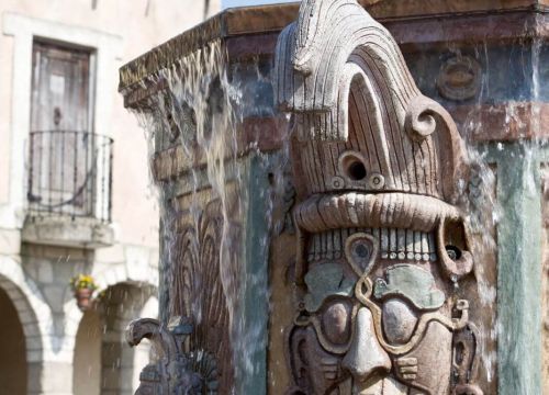 Am Brunnen sind Figuren mit verschiedenen Löchern. Oben kommt aus dem Brunnen Wasser raus.