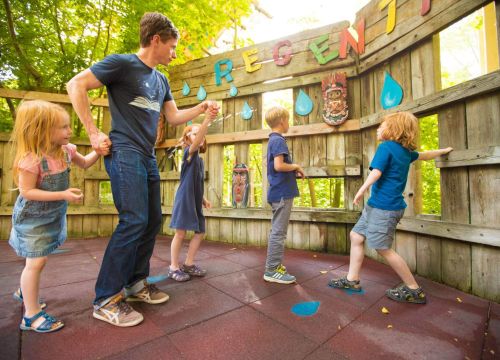 Ein Erwachsener und vier Kinder sind beim Regentanz. Aus einem Loch der Wand kommt ein Wasserstrahl.