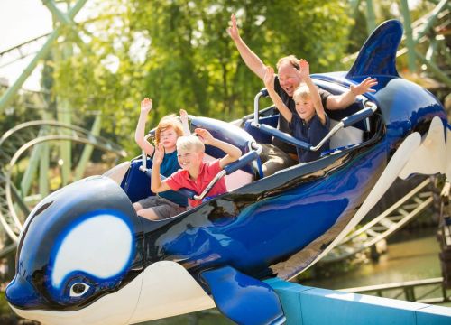 Ein Orca der Attraktion Flying Orca mit vier Gästen drin. Sie freuen sich und haben die Arme oben.