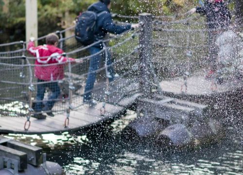 Eine Familie auf der Schwimm- und Wackelbrücke im Hintergrund. Eine Wasserfontäne im Vordergrund.