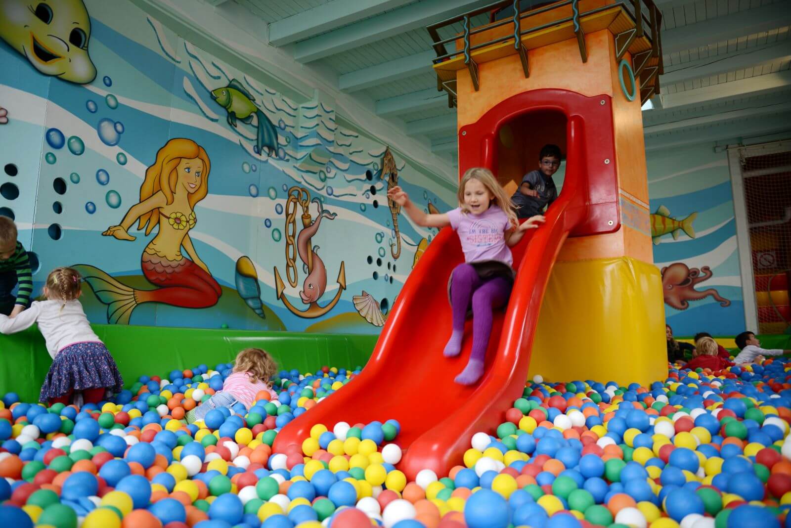 Ganz viele bunte Bälle zwischen den eine Rutsche steht. Ganz viele Kinder sind im Bällebad.