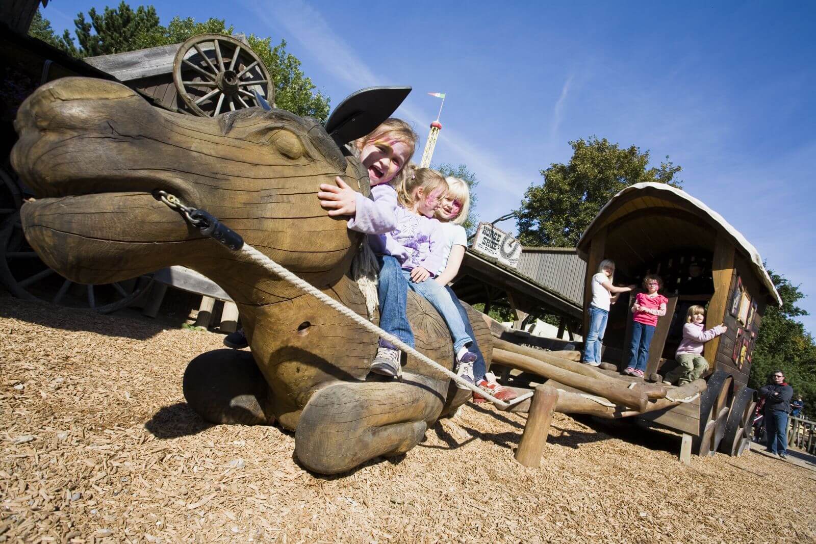 Drei Mädchen sitzen auf einem liegenden Pferd. Auf der Postkutsche dahinter stehen drei Mädchen.