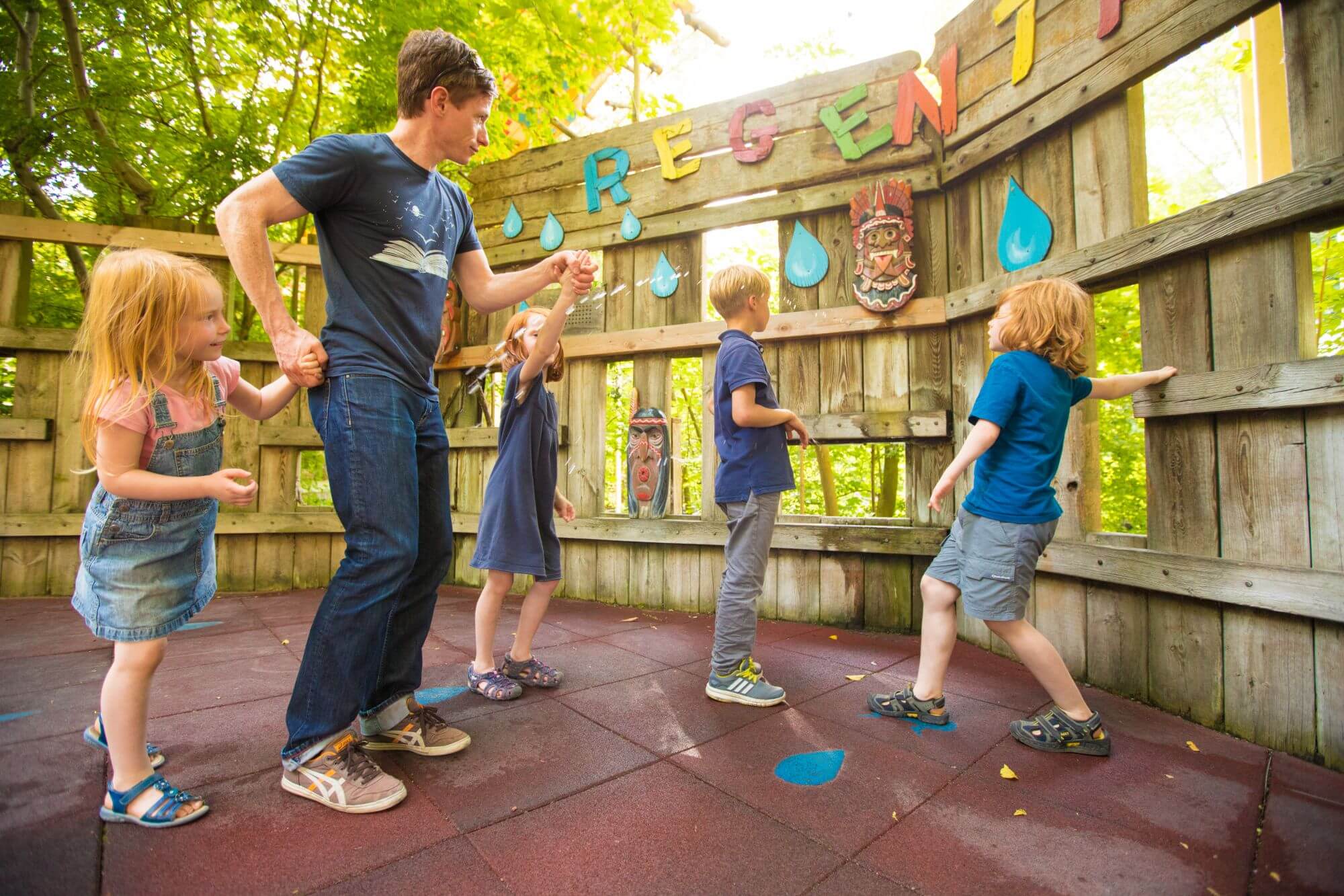 Ein Erwachsener und vier Kinder sind beim Regentanz. Aus einem Loch der Wand kommt ein Wasserstrahl.