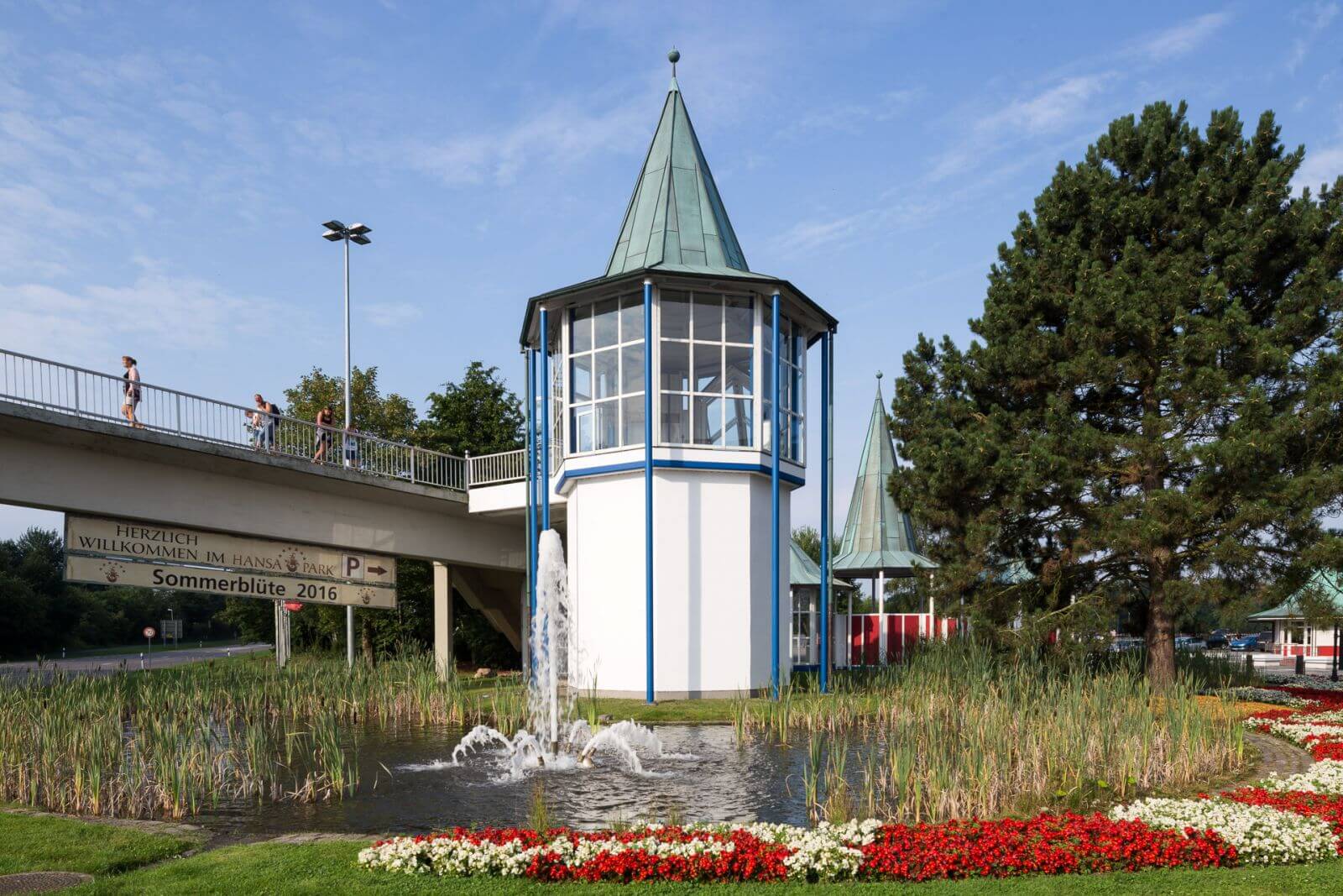Der Rolltreppenturm ist Zentral im Bild. Man sieht Fußgänger über die Brücke zum HANSA-PARK gehen.