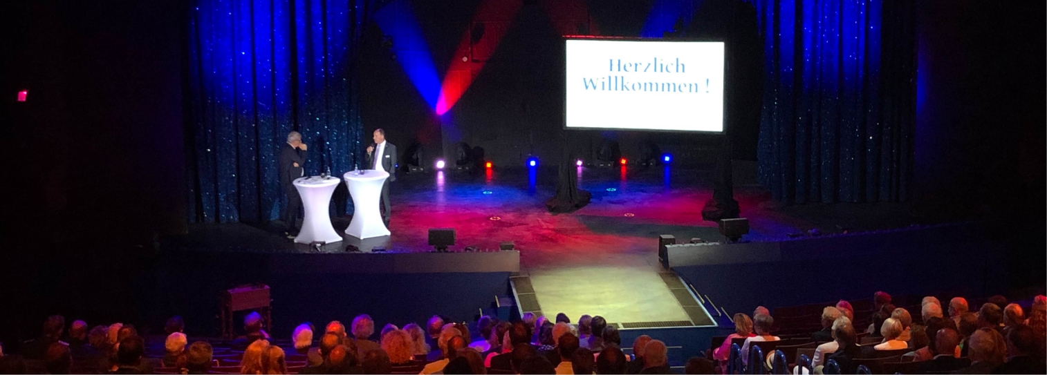 Eine erleuchtete Bühne. Ein Bildschirm zeigt Herzlich Willkommen.