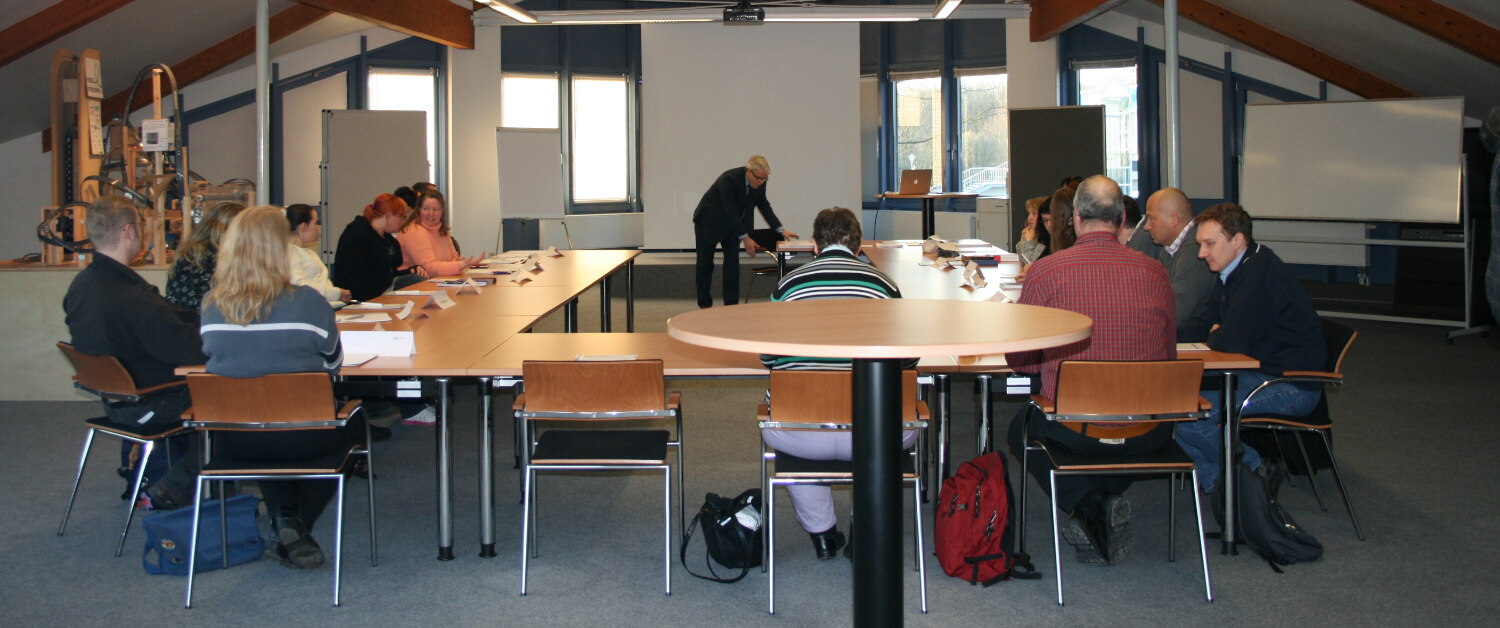 Menschen in einem Seminarraum. Einer steht, die anderen sitzen. Sie hören aufmerksam zu.