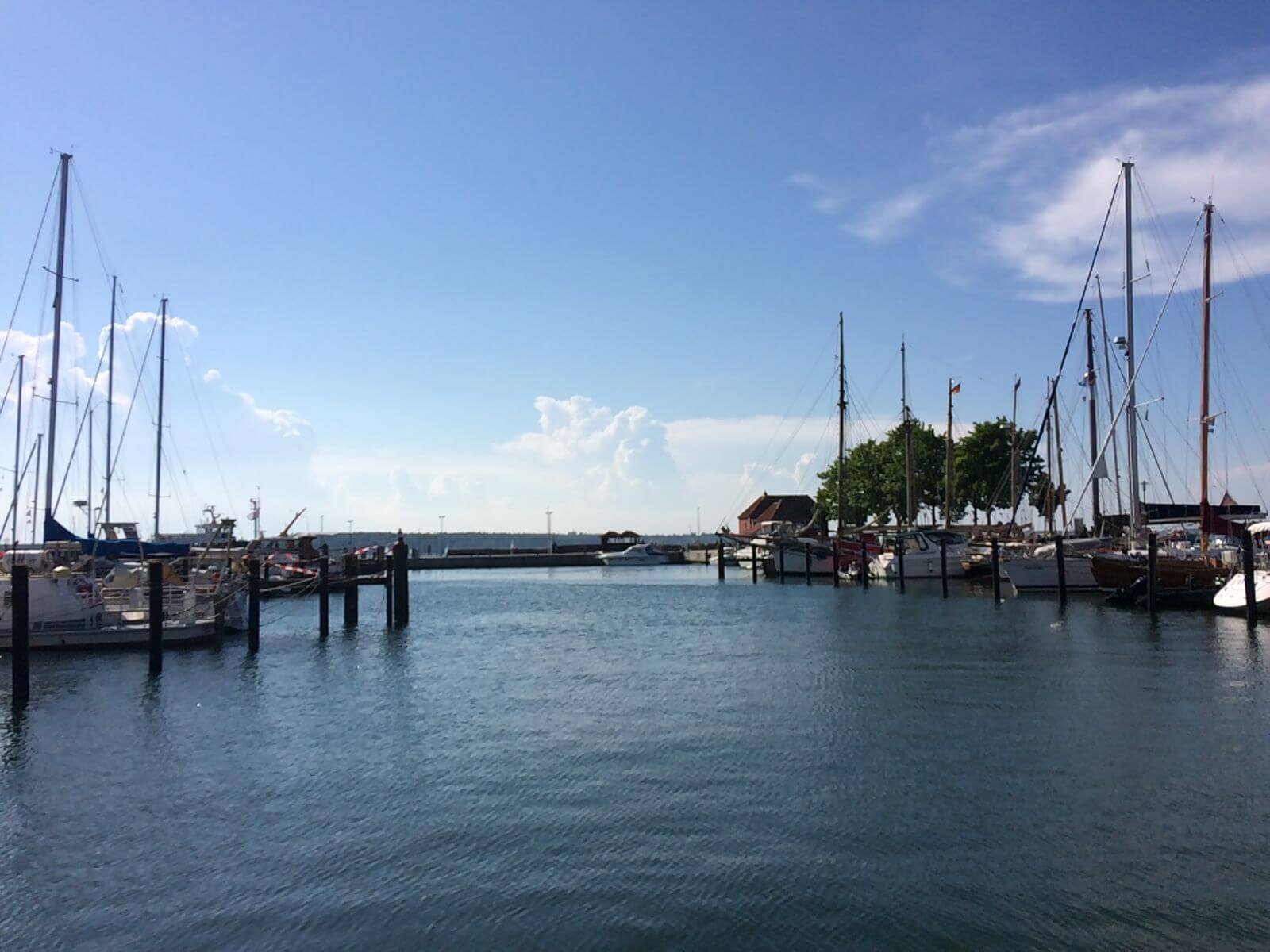 Ein Hafen. Es liegen Segelboote am Hafen an. Der Himmel ist blau.